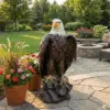 A large, realistic bald eagle statue perched on a rock stands on a stone patio, surrounded by potted flowers and a landscaped garden with trees, lawn, and an outdoor fire pit in the background.