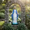 A serene, high-detail photograph of a Virgin Mary "Our Lady of Grace" statue, featuring a vibrant blue mantle and white robes, standing atop a blue-tinted cloud base. The statue is centered within a rustic, hand-laid stone archway (alcove) nestled in a lush "Mary Garden." The scene is surrounded by soft white lilies, delicate blue forget-me-nots, and climbing ivy, all illuminated by warm, natural sunlight filtering through a background of soft-focus garden greenery.