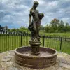A square photo of an ornate, bronze-finished concrete water fountain featuring a standing female figure pouring water from two urns. The intricately carved basin rests on a decorative circular base made of fine pea gravel and weathered flagstones in a residential backyard