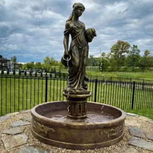 A square photo of an ornate, bronze-finished concrete water fountain featuring a standing female figure pouring water from two urns. The intricately carved basin rests on a decorative circular base made of fine pea gravel and weathered flagstones in a residential backyard